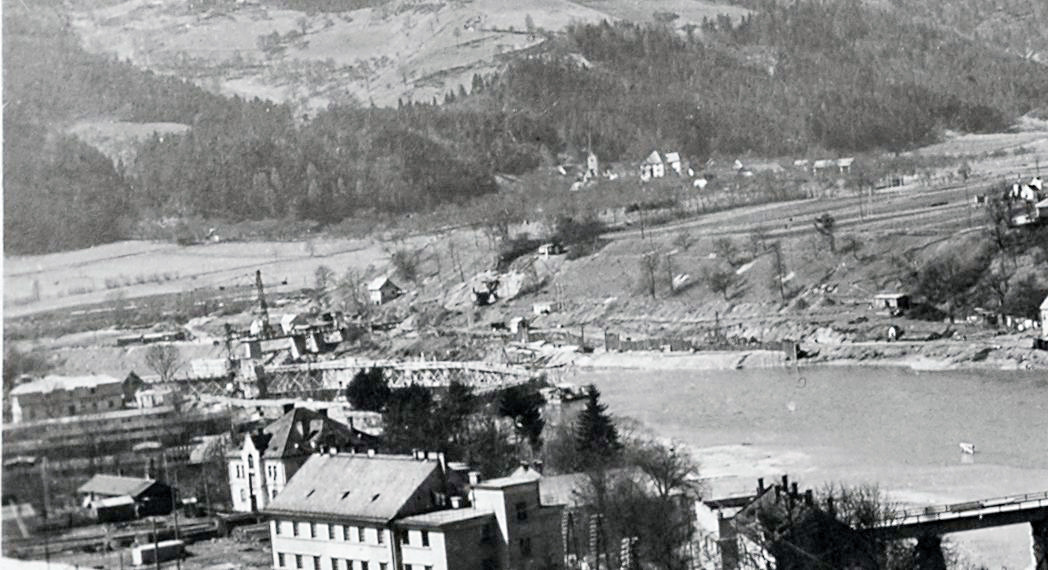 Za Komauerjevim hotelom, železniškim blokom in Gollovo hišo se preko Meže vidi delovno taborišče in gradnja elektrarne. Dravo so preusmerili na levi del struge in na desni gradili prve stebre elektrarne. Takoj zadaj se vidi gradnja novega železniškega mosta, ki so ga, v nasprotju s prepričanji, da je bil stari le popravljen, popolnoma odstranili in zgradili na novo. Vsa polja neposredno za njim so danes zalita z Dravo, vidi se tudi 